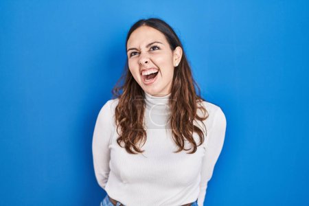 Photo for Young hispanic woman standing over blue background angry and mad screaming frustrated and furious, shouting with anger. rage and aggressive concept. - Royalty Free Image