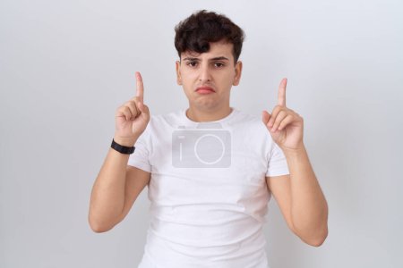 Téléchargez les photos : Jeune homme non binaire portant un t-shirt blanc décontracté pointant vers le haut l'air triste et contrarié, indiquant la direction avec les doigts, malheureux et déprimé. - en image libre de droit