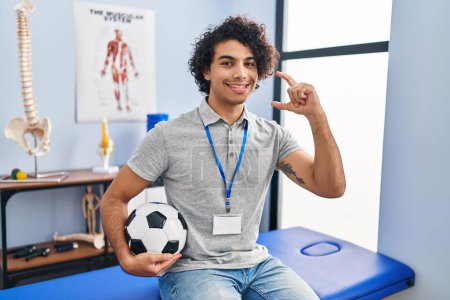 Photo for Hispanic man with curly hair working as football physiotherapist smiling and confident gesturing with hand doing small size sign with fingers looking and the camera. measure concept. - Royalty Free Image