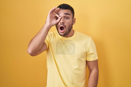 Photo for Young hispanic man standing over yellow background doing ok gesture shocked with surprised face, eye looking through fingers. unbelieving expression. - Royalty Free Image