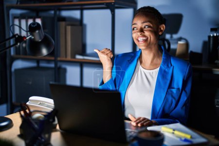 Téléchargez les photos : Belle femme afro-américaine travaillant au bureau la nuit souriant avec le visage heureux regardant et pointant vers le côté avec le pouce vers le haut. - en image libre de droit