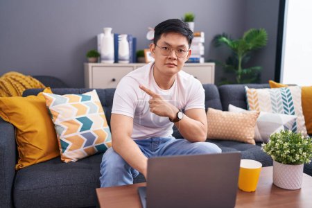 Téléchargez les photos : Jeune homme asiatique à l'aide d'un ordinateur portable à la maison assis sur le canapé pointant avec le doigt de la main sur le côté montrant la publicité, visage sérieux et calme - en image libre de droit