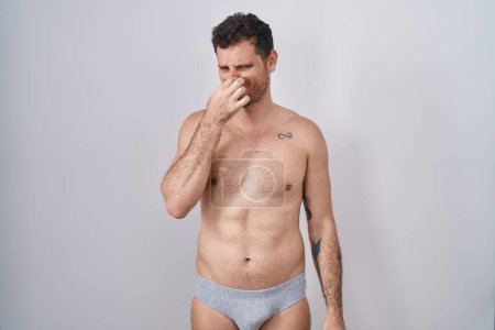 Téléchargez les photos : Jeune homme hispanique debout torse nu portant des sous-vêtements sentant quelque chose de puant et dégoûtant, odeur intolérable, retenant la respiration avec les doigts sur le nez. mauvaise odeur - en image libre de droit