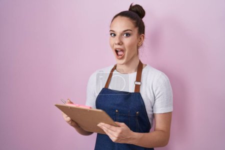 Photo for Young hispanic girl wearing professional waitress apron taking order celebrating crazy and amazed for success with open eyes screaming excited. - Royalty Free Image