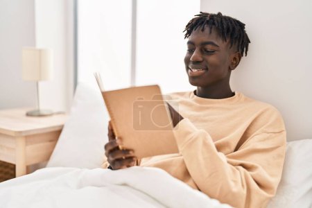 Photo for African american man reading book sitting on bed at bedroom - Royalty Free Image