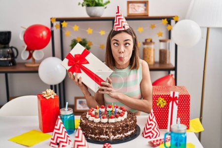 Téléchargez les photos : Femme hispanique célébrant l'anniversaire avec gâteau tenant cadeau faisant visage de poisson avec la bouche et les yeux plissés, fou et comique. - en image libre de droit