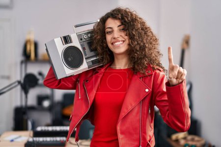 Foto de Mujer hispana con el pelo rizado sosteniendo boombox, escuchando música sonriendo con una idea o pregunta señalando el dedo con la cara feliz, número uno - Imagen libre de derechos