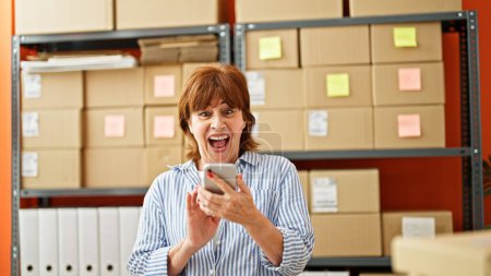 Téléchargez les photos : Femme d'âge moyen e-commerce travailleur d'affaires en utilisant smartphone avec geste gagnant au bureau - en image libre de droit