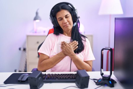 Téléchargez les photos : Femme hispanique mature jouant à des jeux vidéo à la maison souriant avec les mains sur la poitrine avec les yeux fermés et geste reconnaissant sur le visage. concept de santé. - en image libre de droit