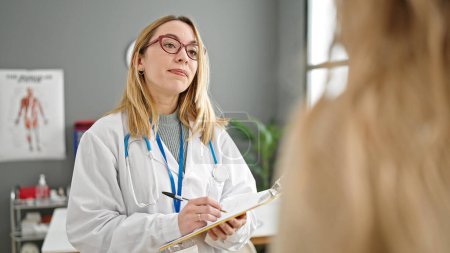 Téléchargez les photos : Jeune femme blonde médecin ayant consultation médicale écrit sur le document à la clinique - en image libre de droit