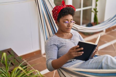 Photo for African american woman reading book lying on hammock at home terrace - Royalty Free Image