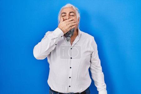 Foto de Hombre de mediana edad con el pelo gris de pie sobre fondo azul aburrido bostezo cansado cubriendo la boca con la mano. inquietud y somnolencia. - Imagen libre de derechos