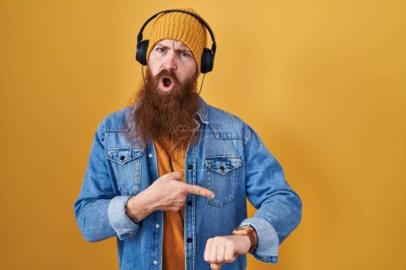 Téléchargez les photos : Homme caucasien à longue barbe écoutant de la musique à l'aide d'écouteurs pressés pointant vers regarder le temps, l'impatience, contrarié et en colère pour retard de délai - en image libre de droit