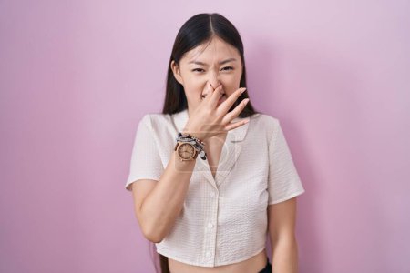 Téléchargez les photos : Jeune femme chinoise debout sur fond rose sentant quelque chose de puant et dégoûtant, odeur intolérable, retenant souffle avec les doigts sur le nez. mauvaise odeur - en image libre de droit