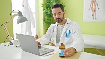 Foto de Joven médico hispano usando portátil sosteniendo botella de medicamento en la clínica - Imagen libre de derechos