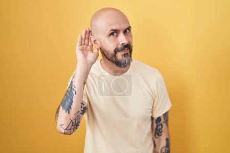 Téléchargez les photos : Homme hispanique avec des tatouages debout sur fond jaune souriant avec la main sur l'oreille en écoutant une rumeur ou des commérages. concept de surdité. - en image libre de droit
