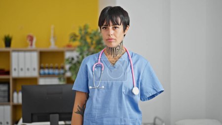 Téléchargez les photos : Femme hispanique avec amputé bras médecin debout avec une expression sérieuse à la clinique - en image libre de droit