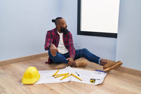 Photo for African american man sitting on the floor at new home looking at blueprints looking to side, relax profile pose with natural face with confident smile. - Royalty Free Image