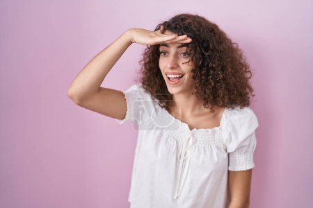 Photo for Hispanic woman with curly hair standing over pink background very happy and smiling looking far away with hand over head. searching concept. - Royalty Free Image