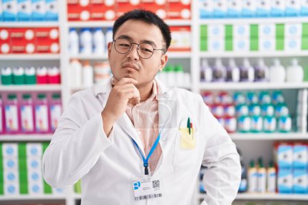 Photo for Chinese young man working at pharmacy drugstore with hand on chin thinking about question, pensive expression. smiling with thoughtful face. doubt concept. - Royalty Free Image