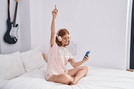 Foto de Young redhead woman listening to music sitting on bed at bedroom - Imagen libre de derechos