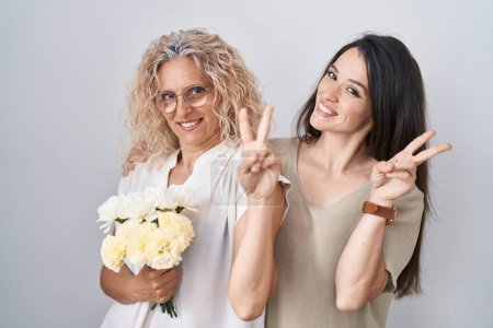 Photo for Mother and daughter holding bouquet of white flowers smiling looking to the camera showing fingers doing victory sign. number two. - Royalty Free Image