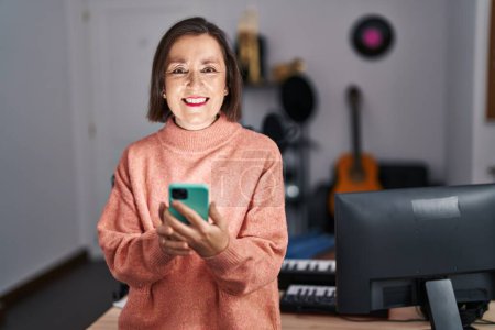 Foto de Mujer de mediana edad músico sonriendo confiado usando teléfono inteligente en el estudio de música - Imagen libre de derechos