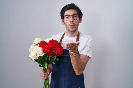 Téléchargez les photos : Jeune homme hispanique tenant bouquet de roses blanches et rouges regardant la caméra souffler un baiser avec la main sur l'air étant belle et sexy. expression d'amour. - en image libre de droit