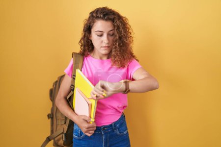 Photo for Young caucasian woman wearing student backpack and holding books checking the time on wrist watch, relaxed and confident - Royalty Free Image