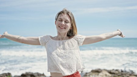 Foto de Joven mujer rubia turista sonriendo confiado de pie con los brazos abiertos en la playa - Imagen libre de derechos