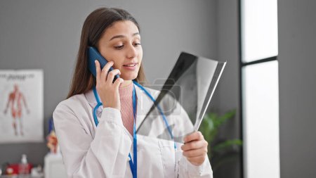 Foto de Joven hermosa mujer hispana médico buscando rayos X hablando en el teléfono inteligente en la clínica - Imagen libre de derechos