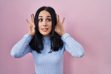 Photo for Hispanic woman standing over pink background trying to hear both hands on ear gesture, curious for gossip. hearing problem, deaf - Royalty Free Image