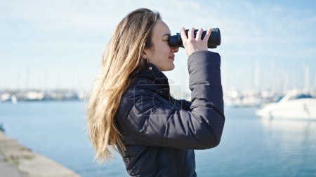 Foto de Joven mujer caucásica sonriendo confiada usando binoculares a la orilla del mar - Imagen libre de derechos