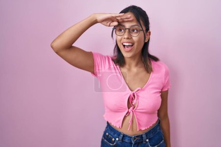 Photo for Hispanic young woman standing over pink background wearing glasses very happy and smiling looking far away with hand over head. searching concept. - Royalty Free Image