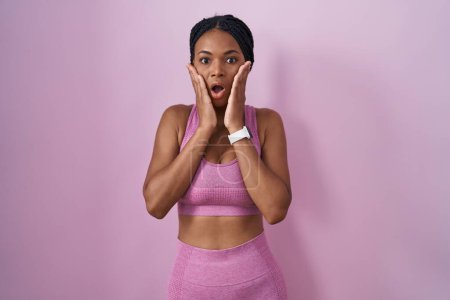 Foto de Mujer afroamericana con trenzas con ropa deportiva sobre fondo rosa asustada y conmocionada, sorpresa y expresión asombrada con las manos en la cara - Imagen libre de derechos