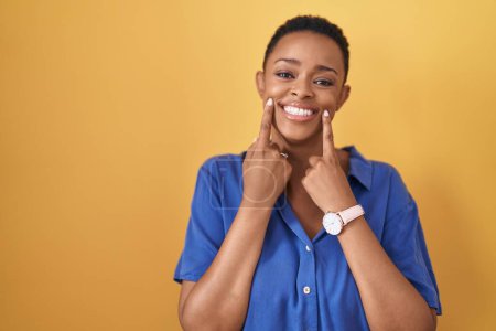 Téléchargez les photos : Femme afro-américaine debout sur fond jaune souriant avec la bouche ouverte, les doigts pointant et forçant sourire joyeux - en image libre de droit