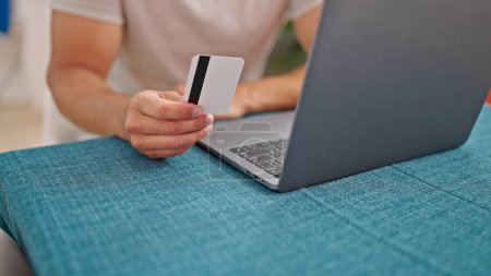Téléchargez les photos : Jeune homme hispanique utilisant un ordinateur portable tenant la carte de crédit assis sur la table à la salle à manger - en image libre de droit
