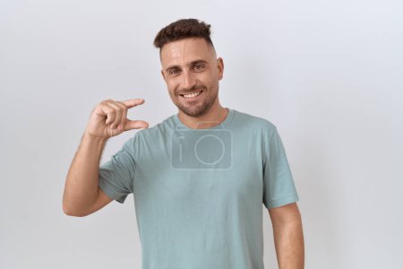 Foto de Hispanic man with beard standing over white background smiling and confident gesturing with hand doing small size sign with fingers looking and the camera. measure concept. - Imagen libre de derechos