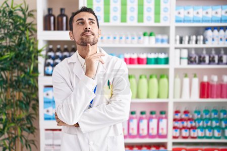 Téléchargez les photos : Bel homme hispanique travaillant à la pharmacie pharmacie pensée concentrée sur le doute avec le doigt sur le menton et la recherche jusqu'à se demander - en image libre de droit