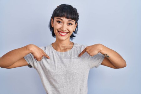Foto de Mujer hispana joven vistiendo camiseta casual sobre fondo azul mirando confiado con sonrisa en la cara, señalándose con los dedos orgullosos y felices. - Imagen libre de derechos