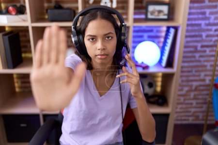 Foto de Joven mujer hispana jugando videojuegos usando auriculares con la mano abierta haciendo stop sign con expresión seria y confiada, gesto de defensa - Imagen libre de derechos