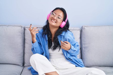 Photo for Young chinese woman listening to music doing guitar gesture at home - Royalty Free Image