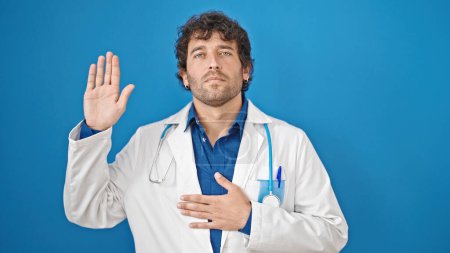 Foto de Joven médico hispano haciendo un juramento con la mano en el pecho sobre un fondo azul aislado - Imagen libre de derechos