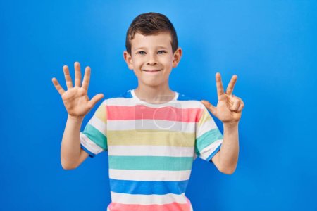 Foto de Joven niño caucásico de pie sobre fondo azul mostrando y apuntando hacia arriba con los dedos número ocho mientras sonríe confiado y feliz. - Imagen libre de derechos