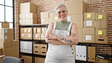Foto de Mujer de pelo gris de mediana edad comercio electrónico trabajador de negocios de pie con los brazos cruzados gesto en la oficina - Imagen libre de derechos