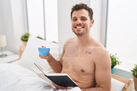 Téléchargez les photos : Jeune homme hispanique lire le livre et boire du café assis sur le lit dans la chambre - en image libre de droit