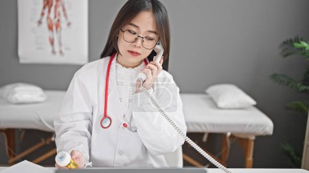Foto de Mujer joven china médico prescribir pastillas tratamiento hablando por teléfono en la clínica - Imagen libre de derechos