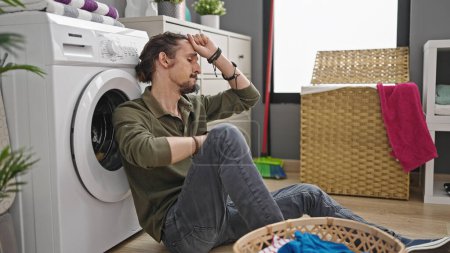 Photo for Young hispanic man washing clothes sitting on floor tired at laundry room - Royalty Free Image