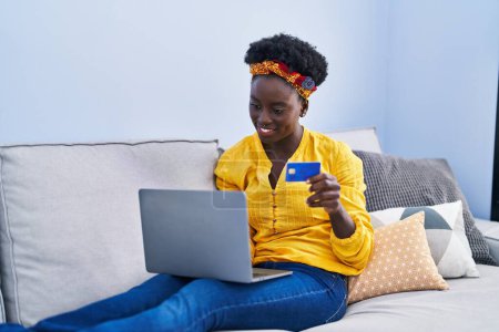 Foto de Mujer afroamericana joven usando portátil y tarjeta de crédito sentado en el sofá en casa - Imagen libre de derechos
