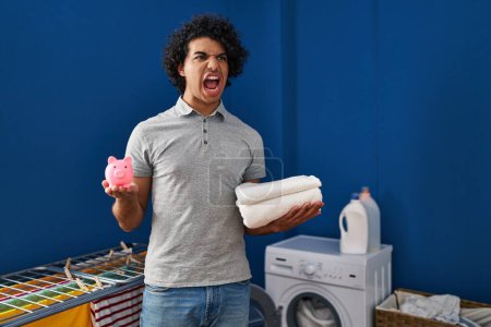 Photo for Hispanic man with curly hair holding clean towels and piggy bank angry and mad screaming frustrated and furious, shouting with anger. rage and aggressive concept. - Royalty Free Image
